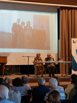 Das Ehepaar Michalski sitzt an einem Tisch auf der Bühne. Im Hintergrund eine alte Fotografie, die auf die Leinwand projiziert wird. 