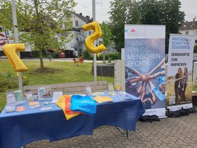 Ein Tisch steht im Freien, darauf eine blaue Tischdecke und verschiedene Materialien, daneben zwei Roll-ups.