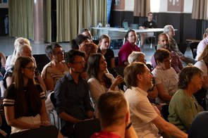 Zuschauende, die sitzen; eine Zuschauende hält ein Mikrofon in der Hand.