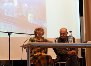 Das Ehepaar Michalski sitzt an einem Tisch auf der Bühne, im Hintergrund ein vergrößertes Foto, wahrscheinlich einer Stadt.