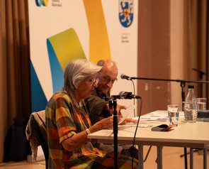 Frau Michalski sitzt am Tisch und hält einen Zettel in der Hand. Daneben sitzt Herr Michalski.