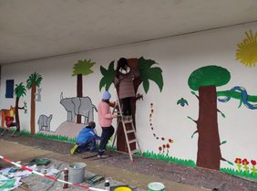 Kinder bemalen die Wand auf dem Schulhof, ein Kind steht auf einer Leiter.