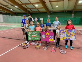 Kinder stehen an einem Tennisnetz und halten Plakate, dahinter stehen auch zwei Erwachsene.
