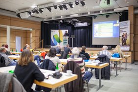 Menschen sitzen an Tischen, auf der Bühne hält ein Referent einen Vortrag.