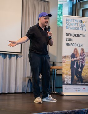 Der Referent Tobias Ginsburg steht auf der Bühne und hält ein Mikrofon in der rechten Hand.