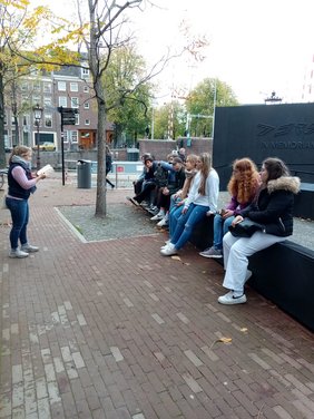 Jugendliche sitzen an einem Denkmal, davor steht eine Person und referiert.