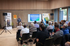 Teilnehmer der Veranstaltung sitzen auf Stühlen, vorne steht ein Referent.