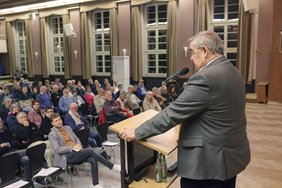 Der Referent Peter Müller steht am Rednerpult und spricht, davor das Publikum auf Stühlen sitzend.