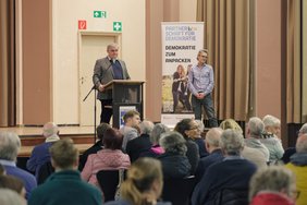 Peter Müller steht am Rednerpult, daneben eine weitere Person, davor das Publikum.