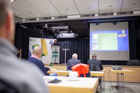 Ein Referent steht auf der Bühne, vor ihm Menschen an Tischen