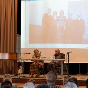Das Ehepaar Michalski sitzt an einem Tisch auf der Bühne, im Hintergrund eine alte Fotografie, die projiziert wird und fünf Menschen zeigt.  