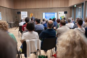 Im Veranstaltungsraum sitzen die Zuschauer auf Stühlen.