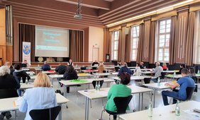 Mehrere Menschen sitzen in einem großen Raum, jeder an einem Tisch, hinten ist zu lesen: Partnerschaft für Demokratie im Landkreis Sankt Wendel.