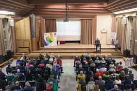 Der Veranstaltungsort aus der Vogelperspektive, vorne eine Bühne mit dem Referenten, davor Stuhlreihen mit Besuchern.