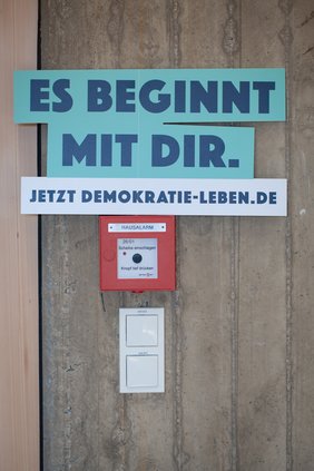 an der wand klebt ein Schild, auf dem steht: Es beginnt mit dir. Jetzt Demokratie leben Punkt DE.