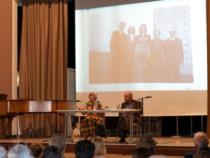 Das Ehepaar Michalski sitzt am Tisch auf der Bühne, im Hintergrund ist eine Fotografie, die fünf Menschen zeigt, zu sehen.