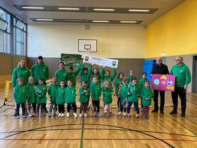 Viele Kinder in grünen Pullovern und einige Erwachsene in einer Halle; hinter ihnen ist ein Tennisnetz.