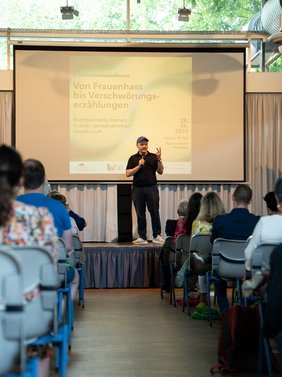 Der Referent Tobias Ginsburg steht auf der Bühne, vor ihm sitzen Zuschauer auf Stühlen.